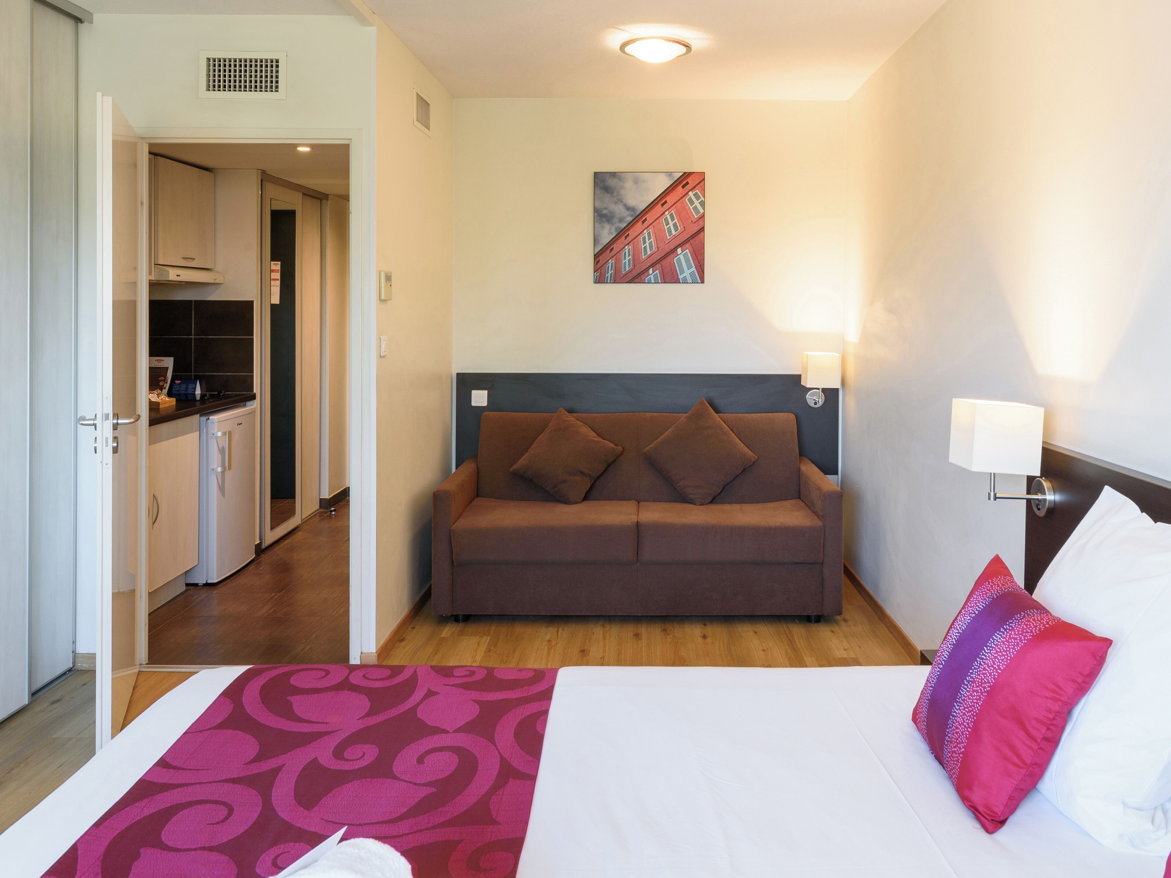 Habitación de hotel con cama, sofá marrón, un cuadro de un edificio, zona de cocina y alfombra estampada.