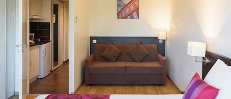 Habitación de hotel con cama, sofá marrón, un cuadro de un edificio, zona de cocina y alfombra estampada.