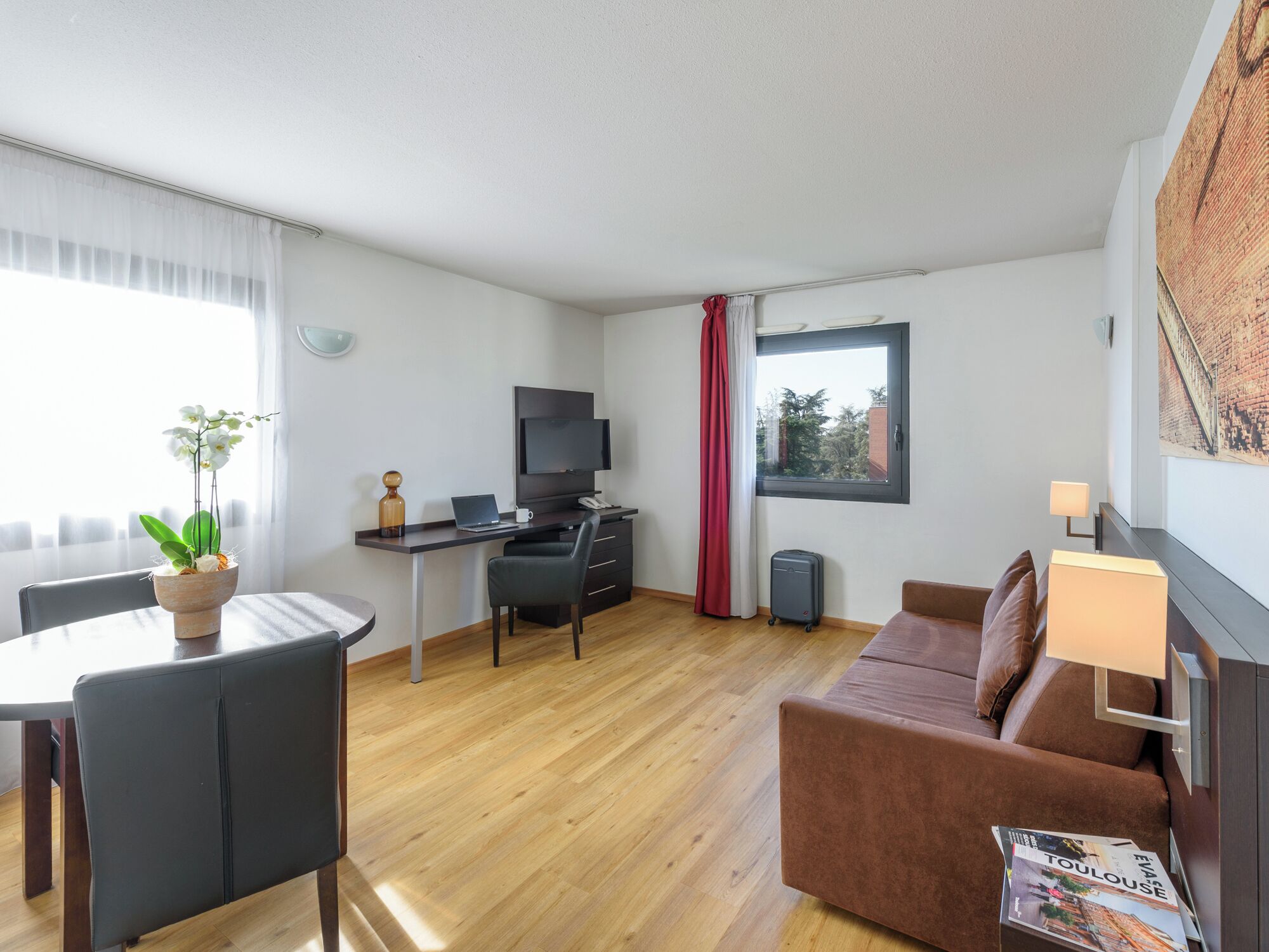 A modern hotel room with a wooden floor, a brown sofa, a dining table with chairs, a desk with a laptop, a TV, and a window with a view of trees.