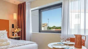 A hotel room with a bed, bedside table with a lamp, a window with curtains, a dining table with a pitcher, and a view of the outside.