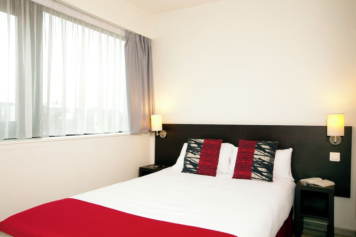 A hotel room with a bed, two bedside tables, a window with curtains, and wall-mounted lamps.