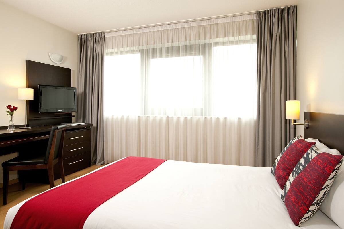 A hotel room with a bed, a desk with a television, a chair, and a window with curtains.