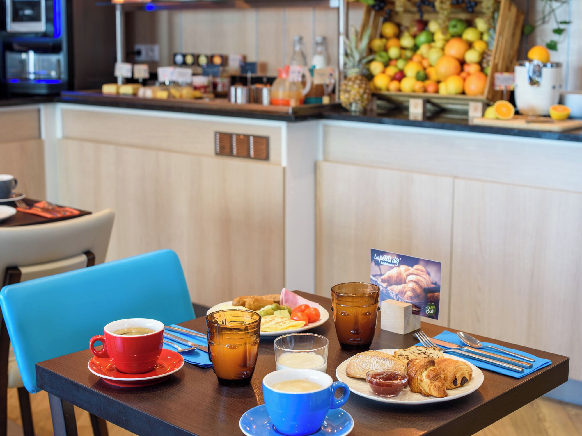 Una mesa de desayuno con café, té, croissants y una presentación de frutas al fondo.