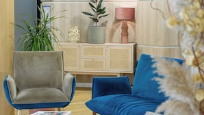 A modern reception area with a wooden reception desk, a blue sofa, and a grey armchair. There is a potted plant and a lamp on the desk.