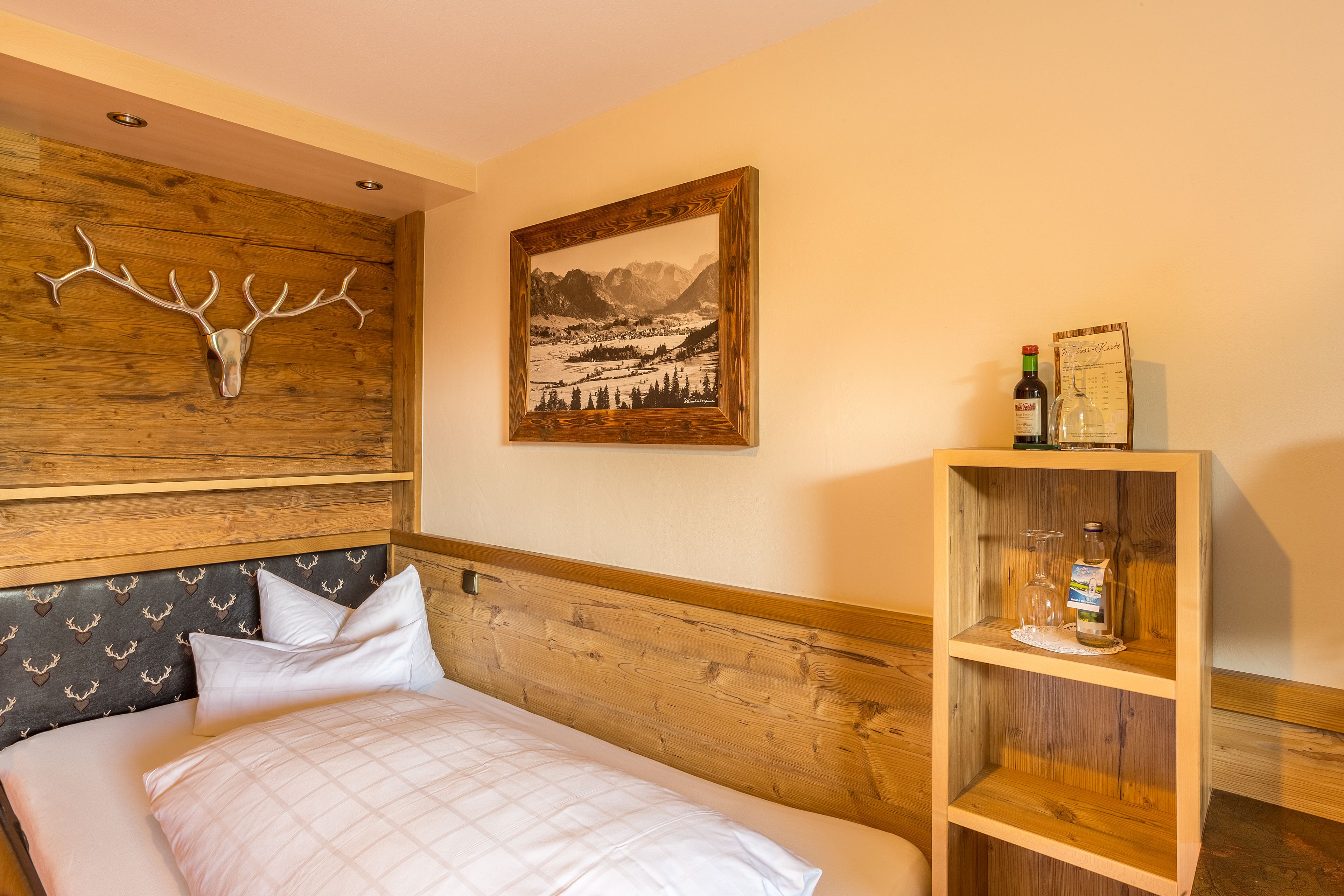 A bedroom with a wooden headboard, a bed with white bedding, a wooden shelf with bottles, and a framed landscape painting.
