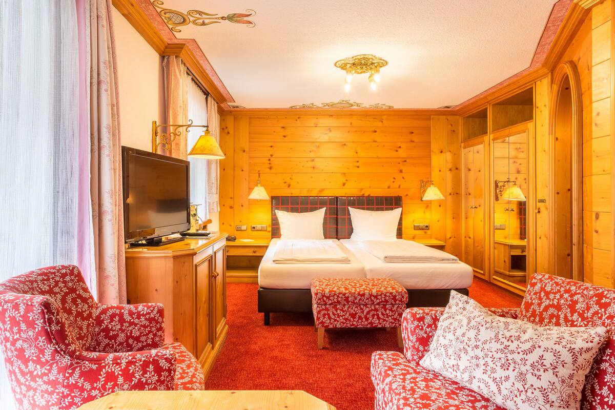 A hotel room with a wooden interior, a bed with white linens, two bedside lamps, a television, and floral-patterned furniture.