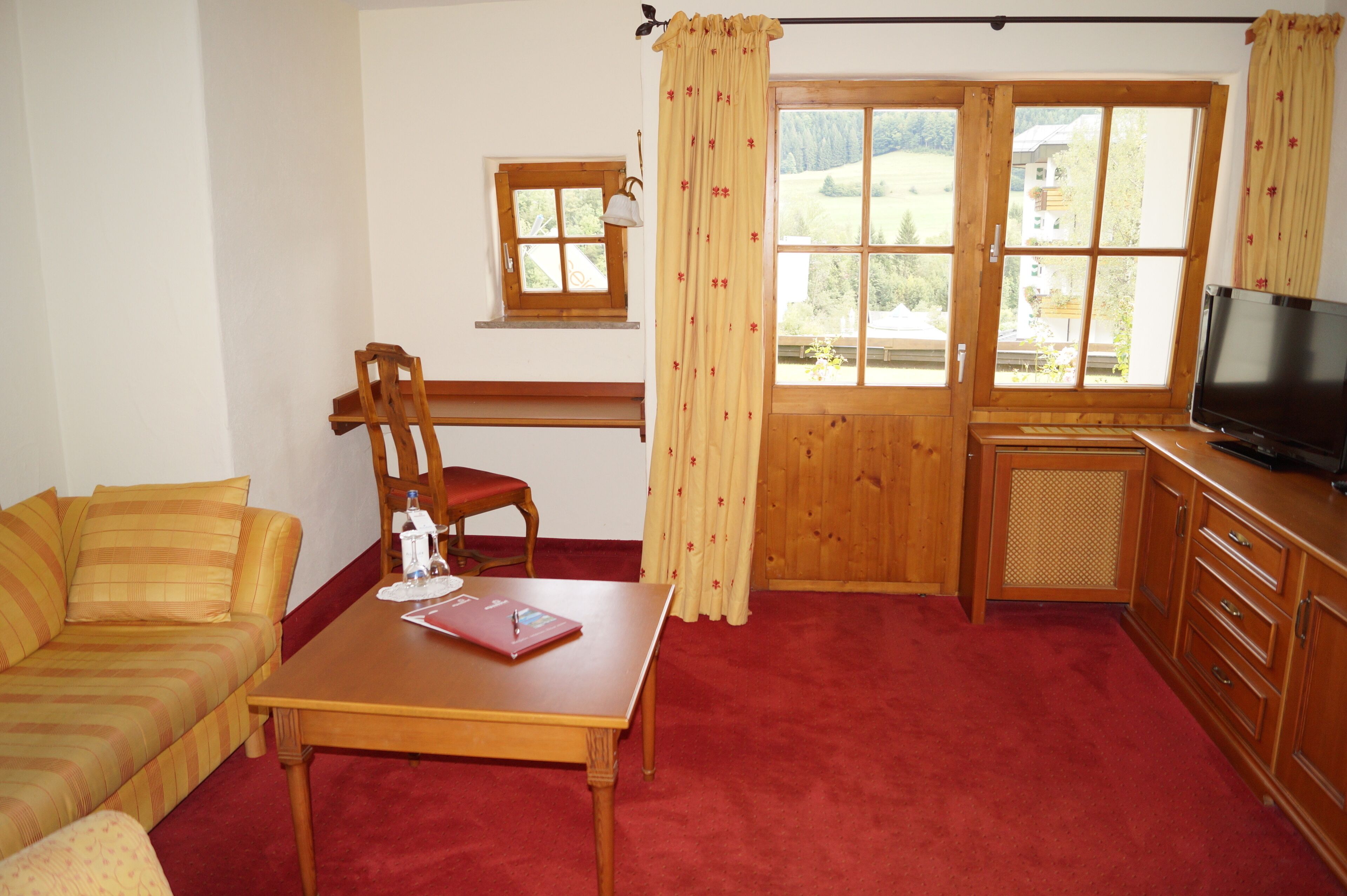 Una sala de estar acogedora con una mesa de madera, un sofá rojo, una silla de madera, un ventanal pequeño y un televisor.