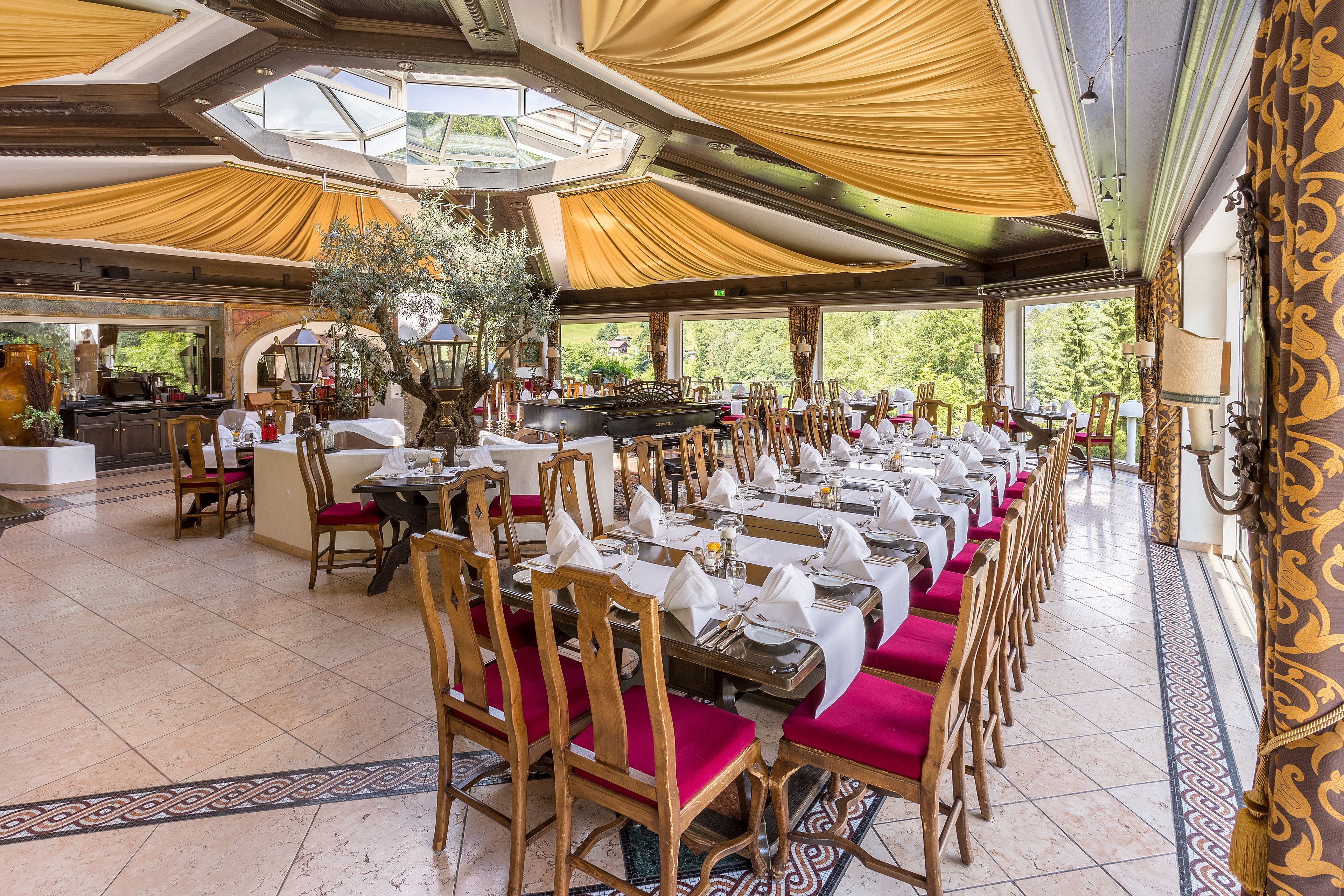 A long dining table with red upholstered chairs, a large chandelier, and a view of the outdoors.