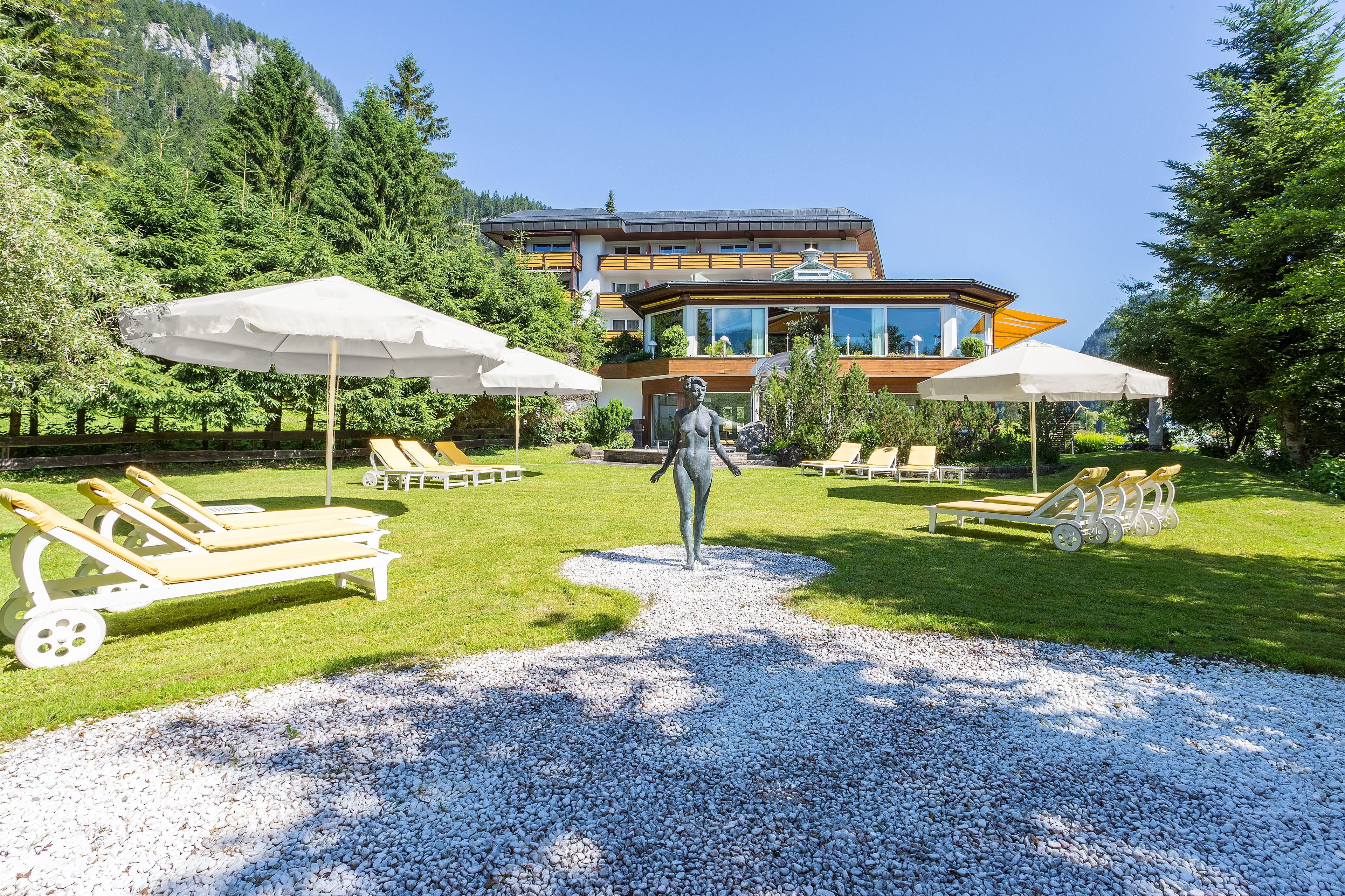 A statue in a garden with lounge chairs and umbrellas, a building in the background, and a mountainous landscape.