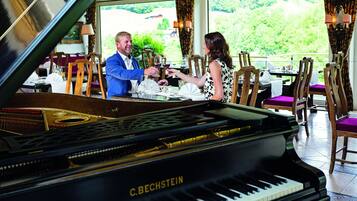 Un piano de cola con la etiqueta C. BECHSTEIN en un comedor con sillas de madera y una mesa dispuesta para dos.
