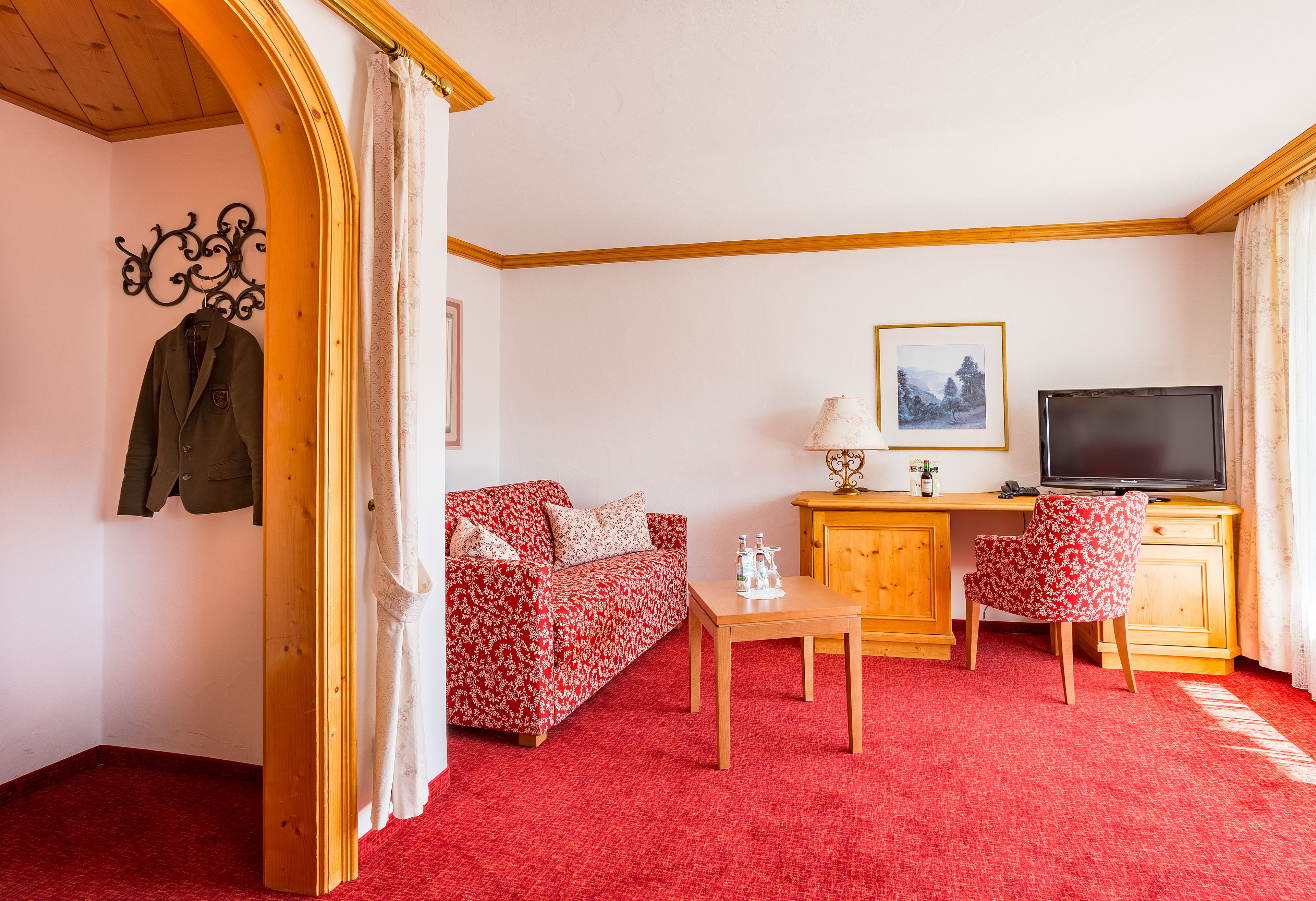 A room with a red carpet, a wooden desk with a TV, a floral sofa, and a coat rack with a jacket.