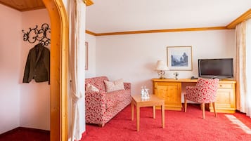 A room with a red carpet, a wooden desk with a TV, a floral sofa, and a coat rack with a jacket.