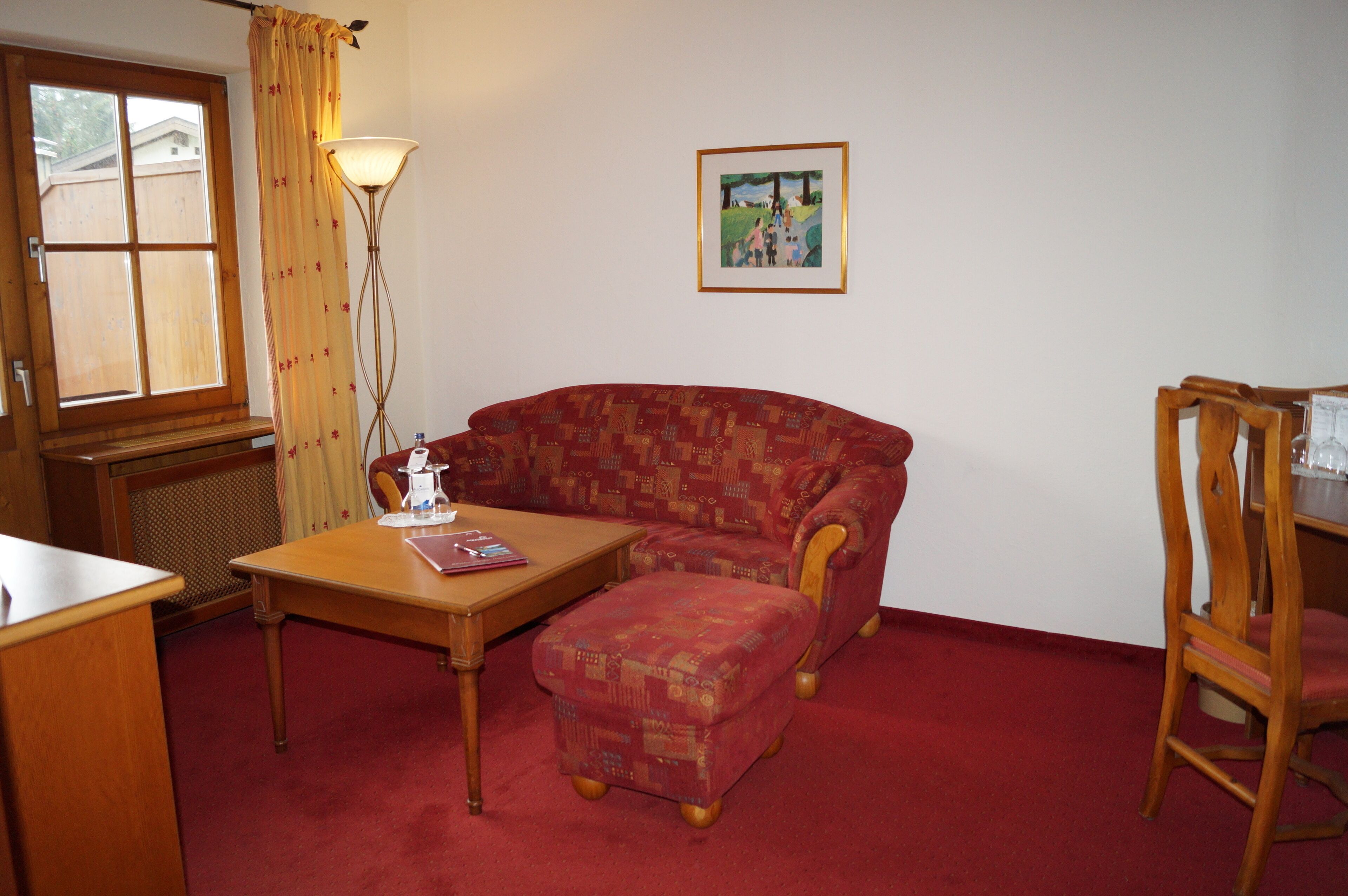 Una habitación con un sofá rojo estampado, una mesa de madera, una lámpara, un cuadro en la pared y una ventana con cortinas.