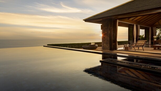 A modern infinity pool with a view of the ocean, a covered outdoor seating area, and a stone facade.