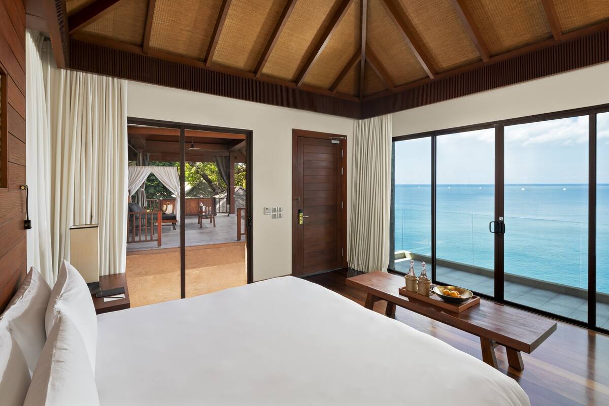 Quarto de hotel com uma cama grande, um banco de madeira com comodidades e uma porta de vidro deslizante para uma varanda com vista para o oceano.