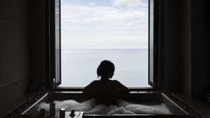 A person sitting in a bathtub with a view of the sea through a large window.