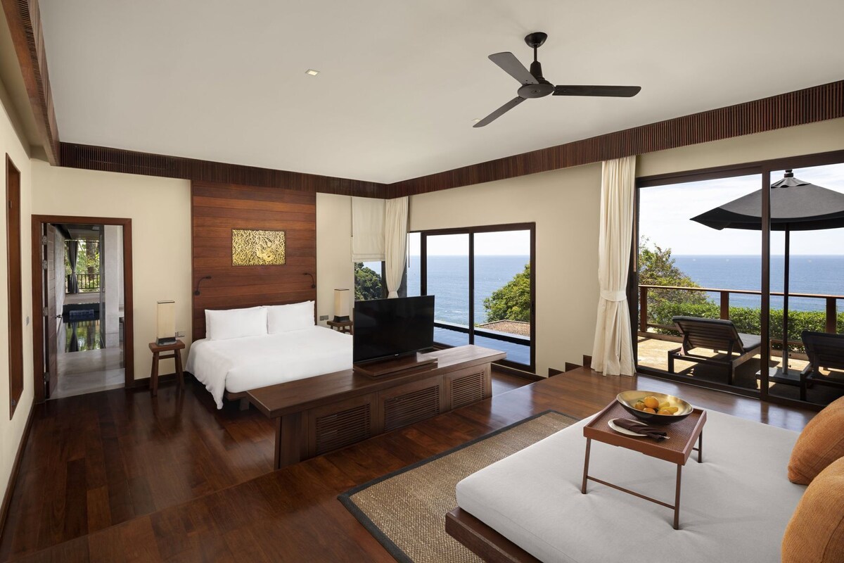 A hotel room with a bed, TV, wooden floor, large windows, and a balcony with outdoor furniture.