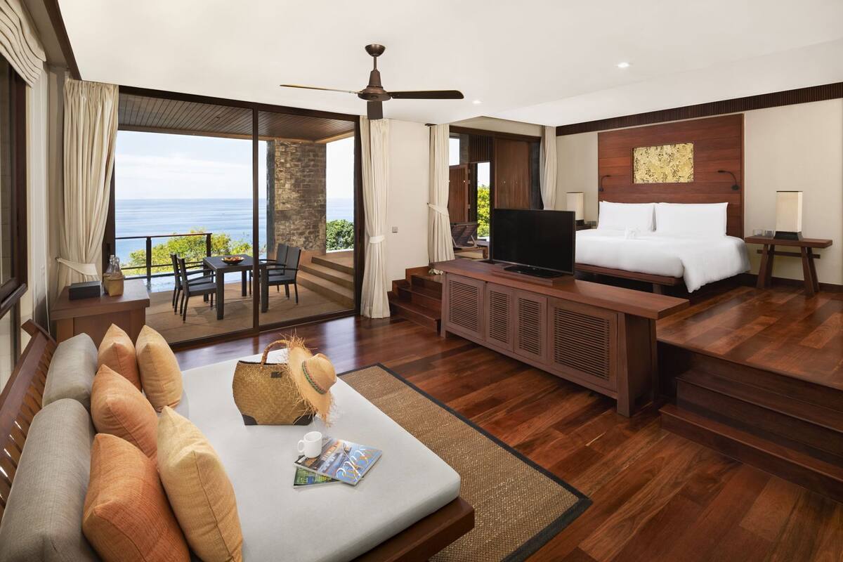 Quarto de hotel com cama grande, televisão, sofá e área de jantar com vista para o mar.