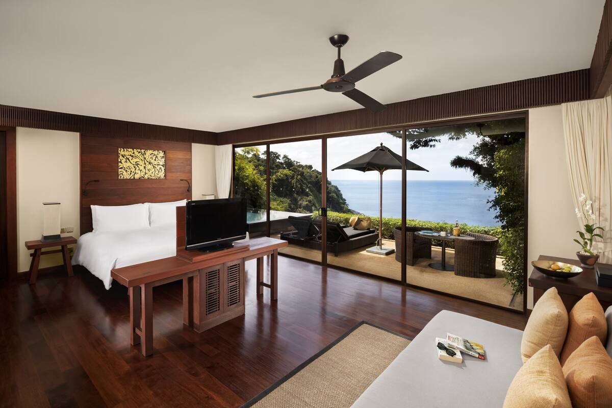 Quarto com uma cama grande, televisão, escrivaninha de madeira e vista para o oceano através de grandes portas de vidro.