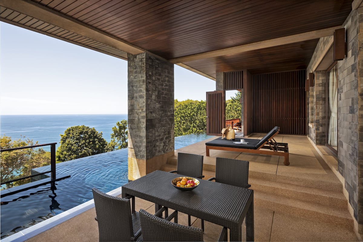 Uma área externa com mesas e cadeiras, vista para o oceano, piscina e paredes de pedra.