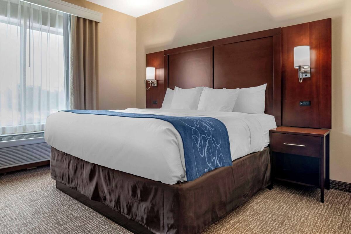 A hotel room with a large bed, a nightstand, a wall-mounted lamp, and a window with curtains.
