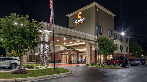 A Comfort Suites hotel with a modern design, featuring a well-lit entrance, a flagpole with an American flag, and a parking lot with several vehicles.