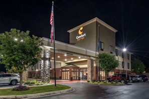 A Comfort Suites hotel with a modern design, featuring a well-lit entrance, a flagpole with an American flag, and a parking lot with several vehicles. - Comfort Suites  Pell CIty near Logan Martin Lake (Pell City)