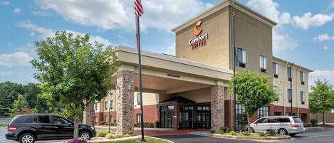 Un hotel Comfort Suites con fachada de ladrillo, una bandera y dos coches aparcados en el estacionamiento.