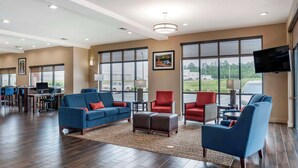 A spacious hotel lobby with a modern design, featuring a large seating area with sofas and armchairs, a flat-screen TV mounted on the wall, and large windows providing views of the outdoors.