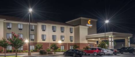 A hotel with a parking lot, multiple cars, and a well-lit exterior.