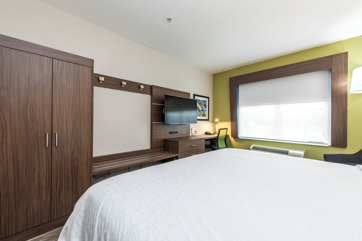 A hotel room with a large bed, a TV mounted on a wooden cabinet, a desk with a chair, and a window with blinds.