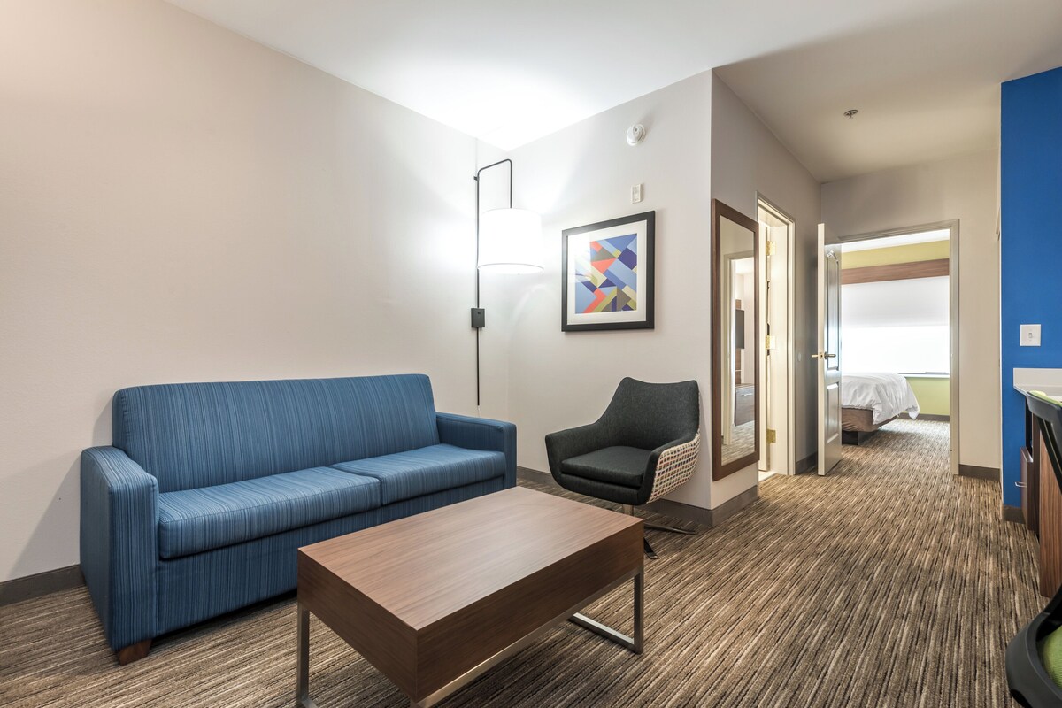 A hotel room with a blue sofa, a wooden coffee table, a black armchair, and a framed abstract art piece on the wall.
