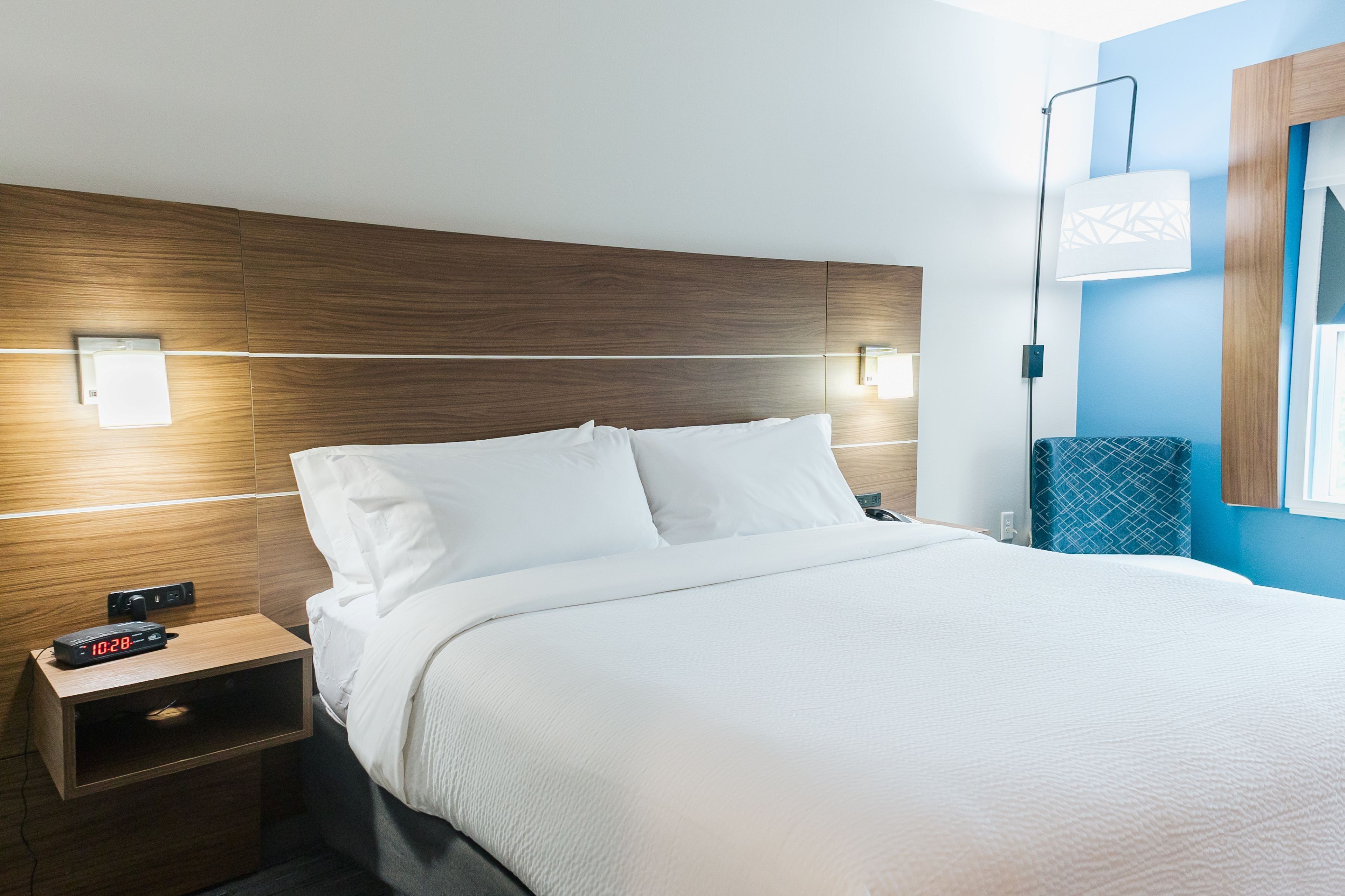 A hotel room with a wooden headboard, a bed with white linens, a bedside table with a digital clock, and a wall-mounted light fixture.