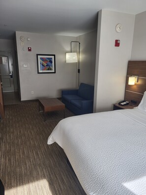 A hotel room with a bed, a blue sofa, a small table, a wall-mounted lamp, and a framed picture.