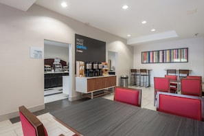 A modern breakfast area with a coffee station, seating, and a sign indicating complimentary breakfast hours.
