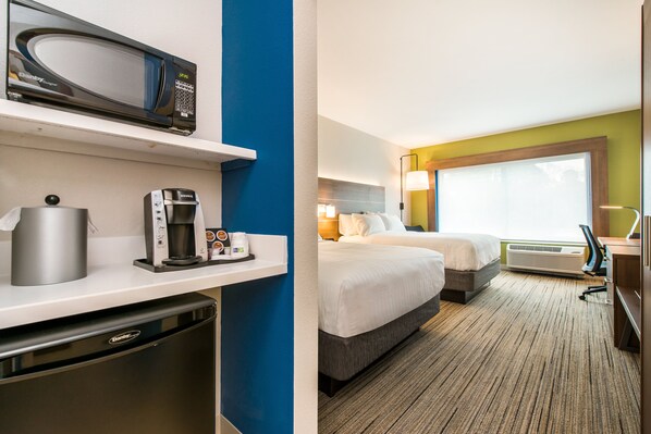 A hotel room with a microwave, coffee maker, and a compact refrigerator. There are two beds with a large window and an air conditioning unit.
