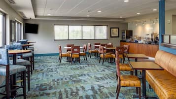 A hotel dining area with multiple tables and chairs, a large window with blinds, a flat-screen TV mounted on the wall, and a kitchen counter with various appliances.