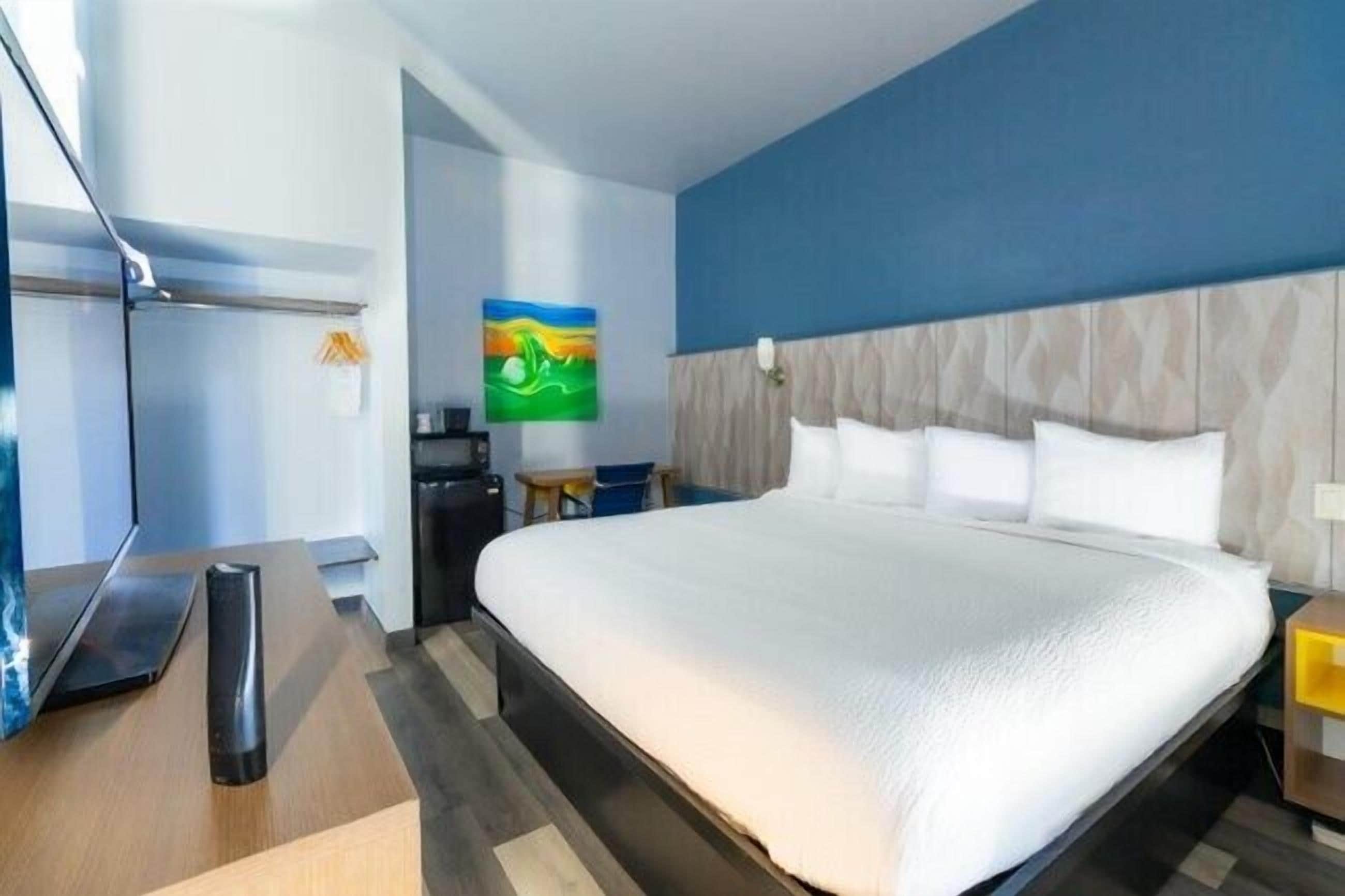 A hotel room with a large bed, a desk with a laptop, a TV, and a painting on the wall.