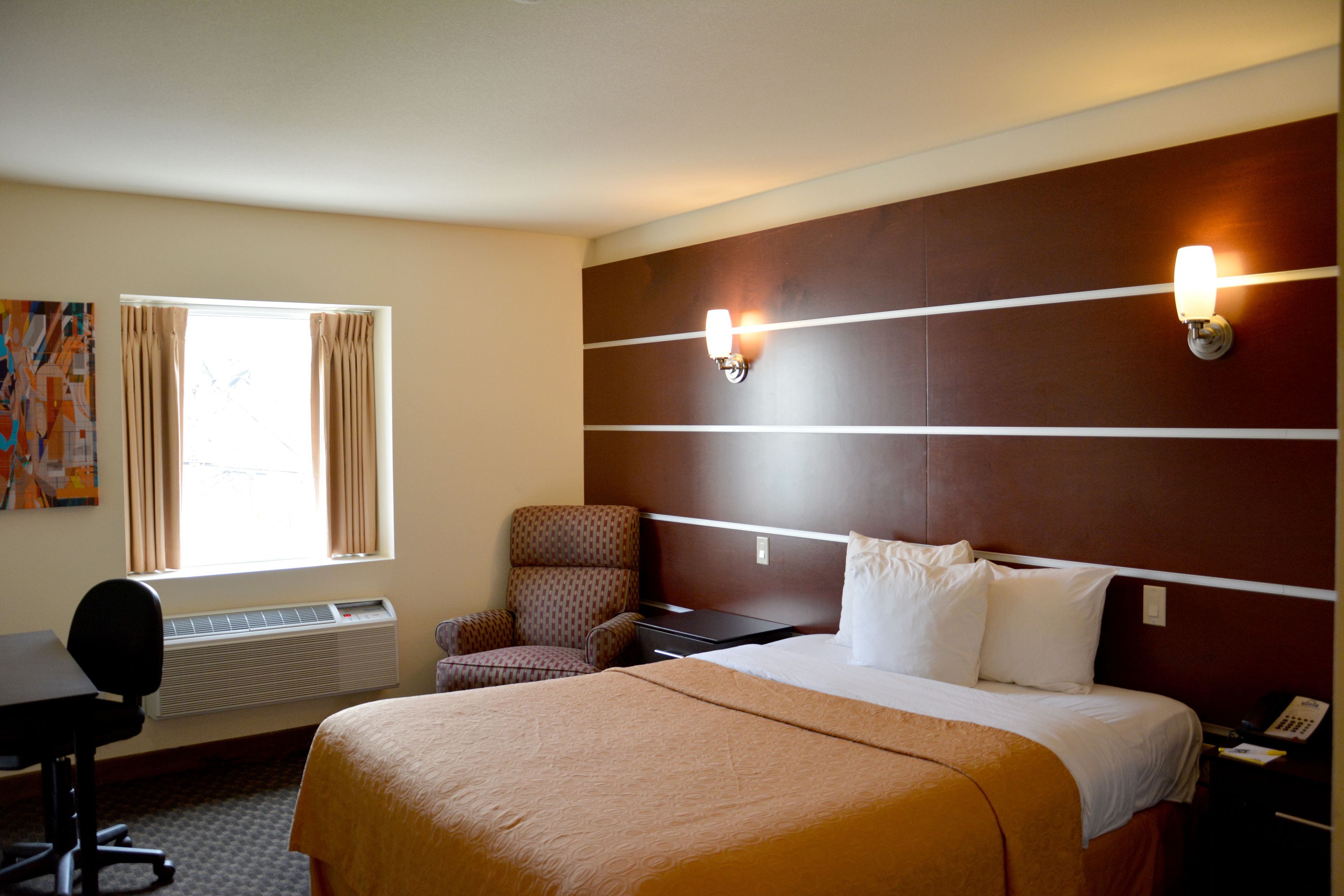 A hotel room with a large bed, a desk with a chair, a wall-mounted phone, and a window with curtains.