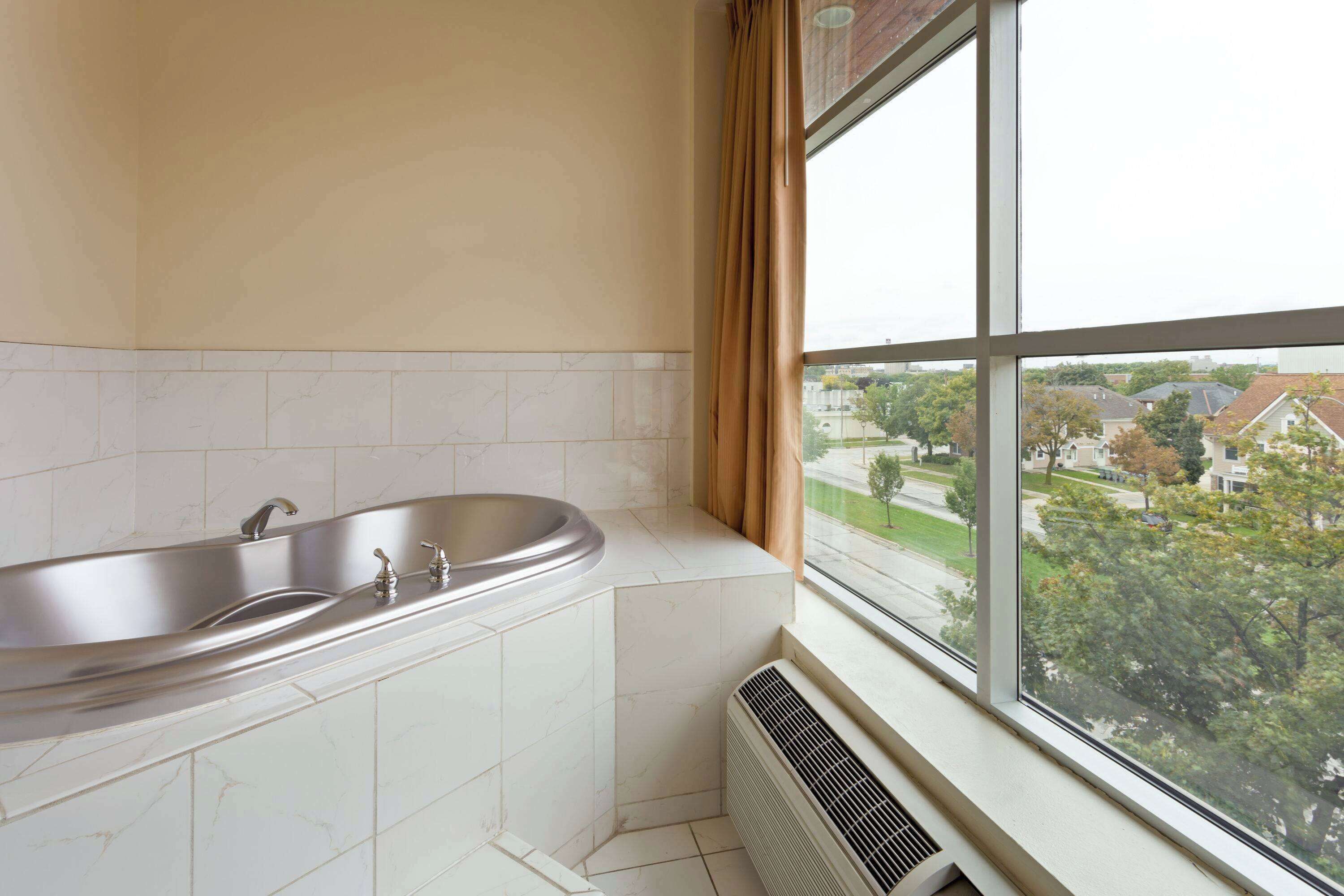 A bathroom with a bathtub, a window with a view of a residential area, and a vent near the window.