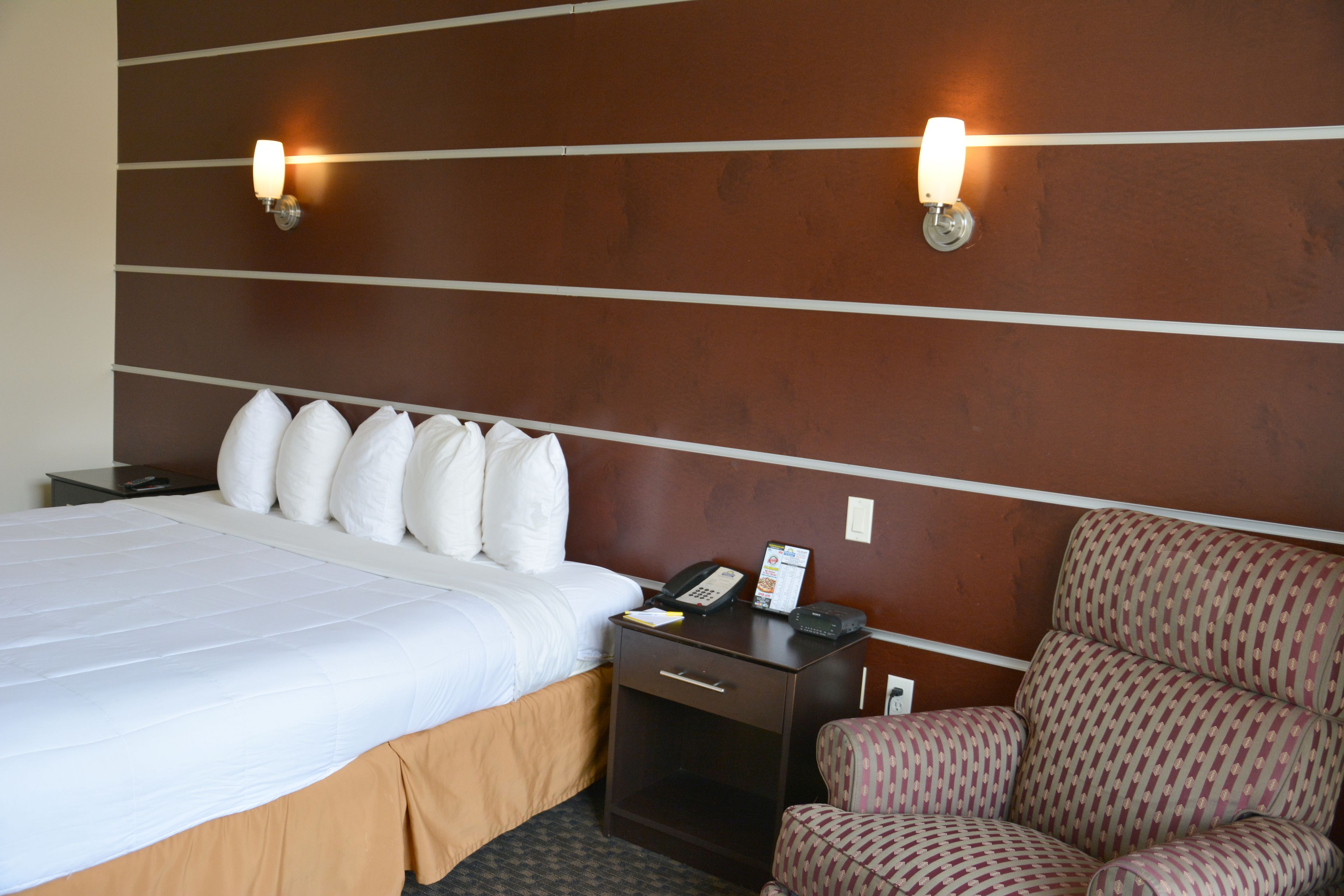 A hotel room with a large bed, a nightstand with a phone and a lamp, and a patterned armchair.