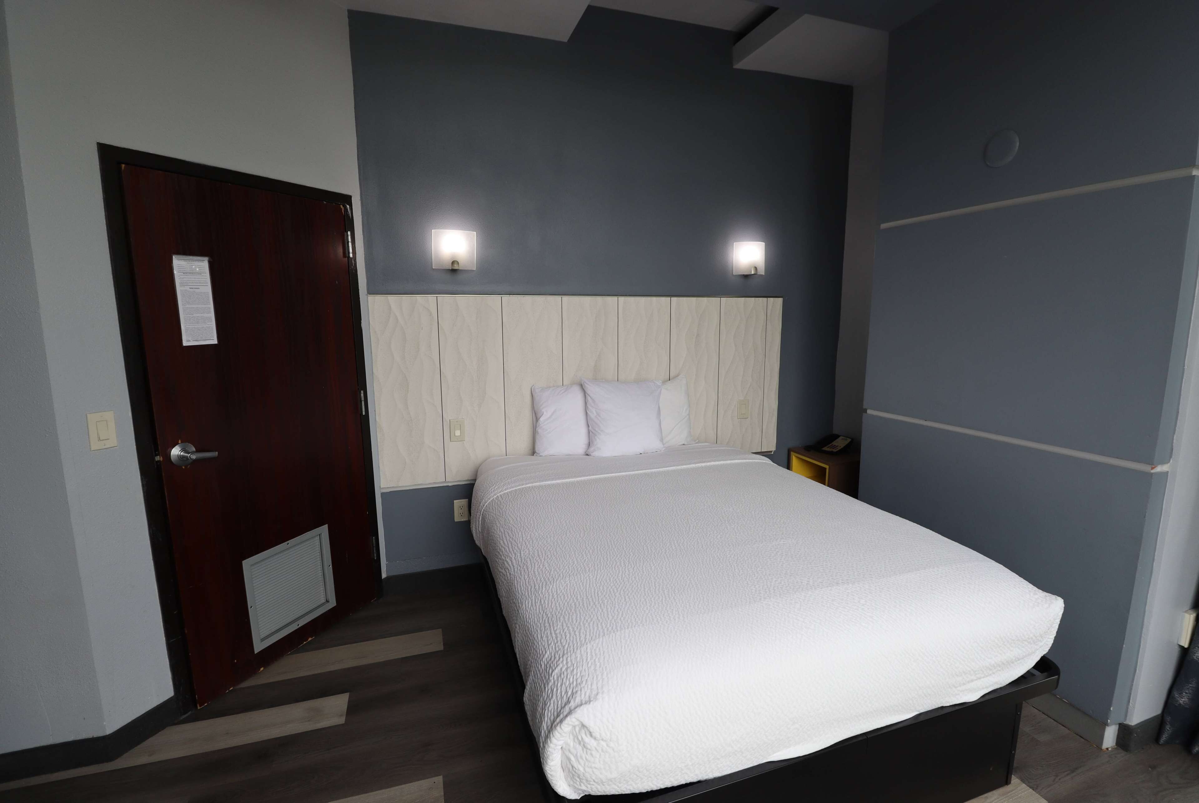 A neatly made bed with a white comforter and pillows, a wooden headboard, and two wall-mounted lights.