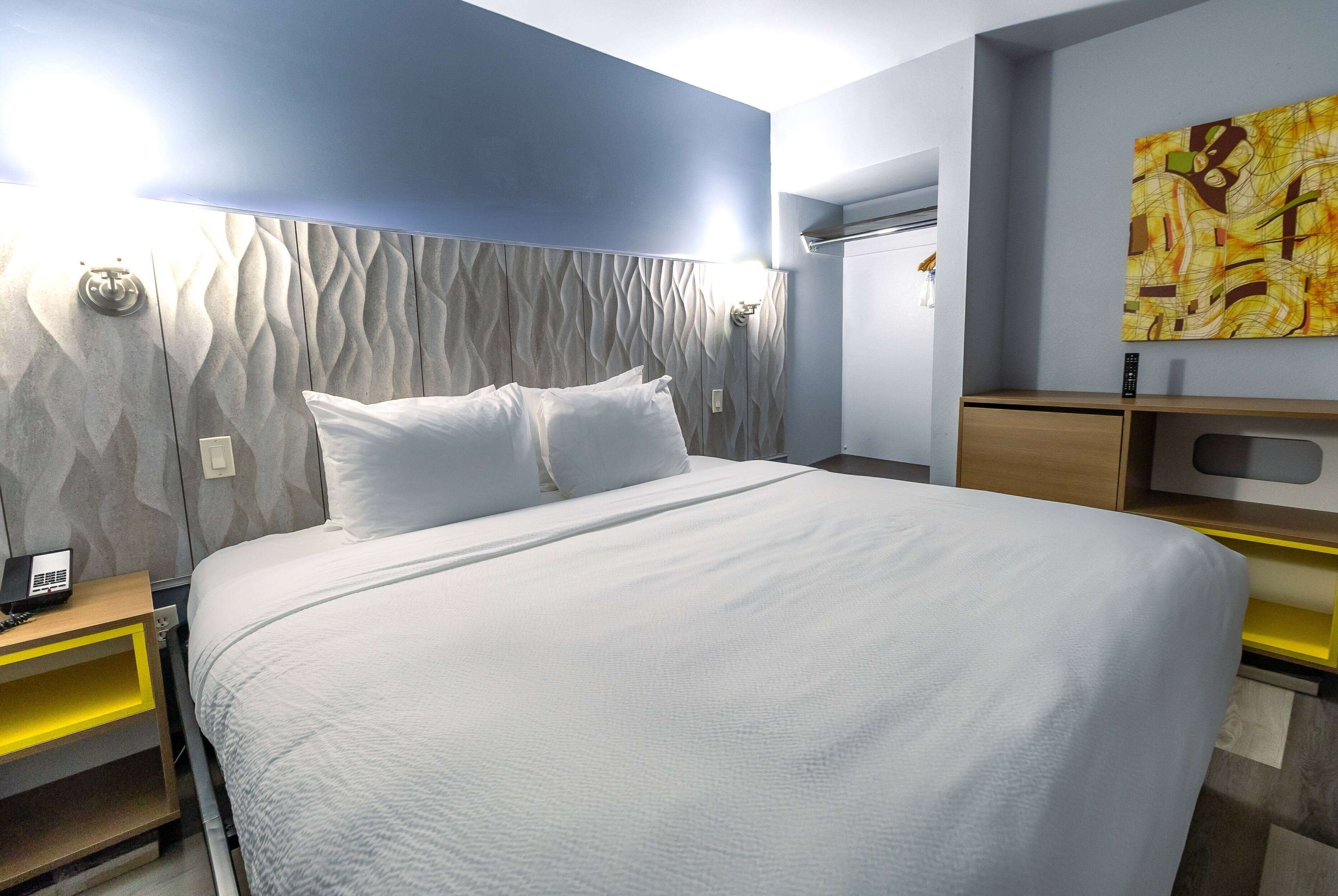 A modern hotel room with a large bed, a nightstand with a phone, a wall-mounted air conditioner, and a painting.