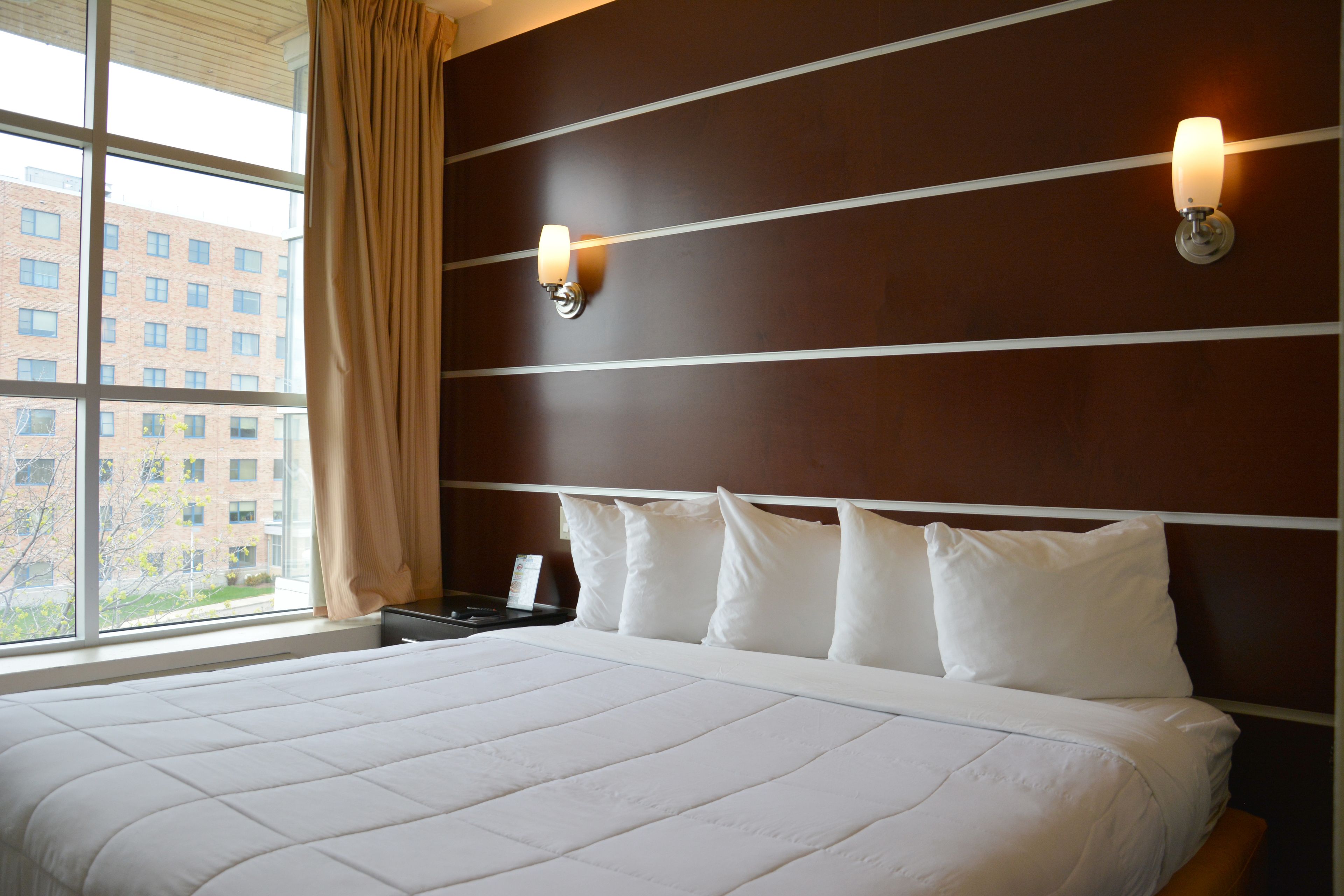 Ein Hotelzimmer mit einem großen Bett, einem Nachttisch, zwei Wandleuchten und einem Fenster mit Vorhängen.