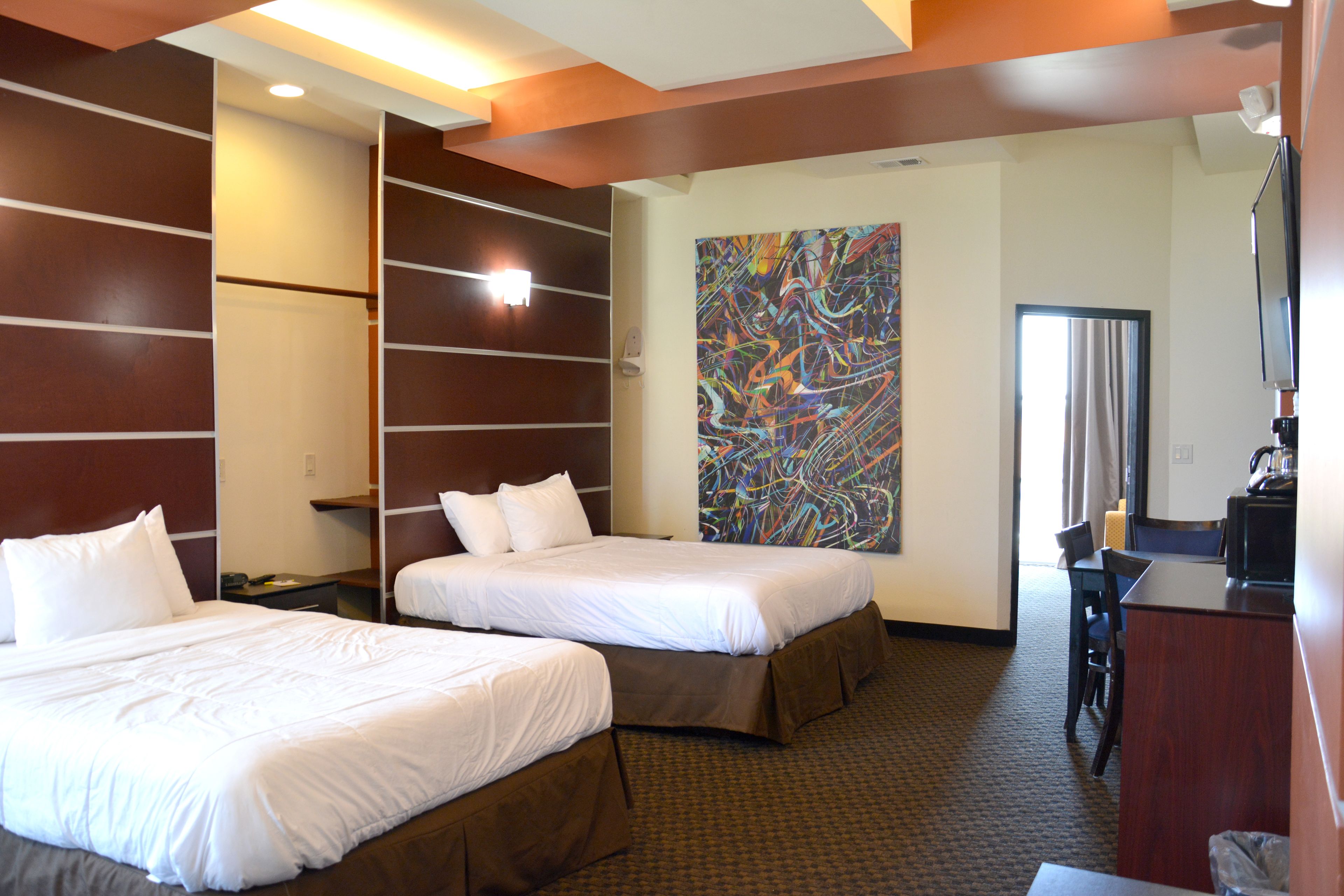Quarto de hotel com duas camas, uma escrivaninha, uma cadeira e um quadro grande e abstrato na parede.