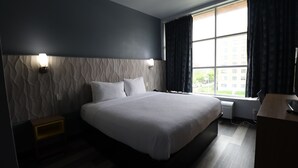 Habitación de hotel moderna con una cama grande, una mesita de noche, un escritorio y un ventanal con cortinas.