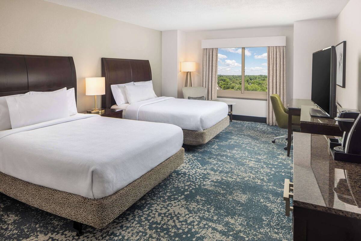 A hotel room with two beds, a desk, a television, and a window with a view of greenery.