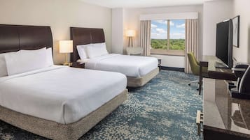 A hotel room with two beds, a desk, a television, and a window with a view of greenery.