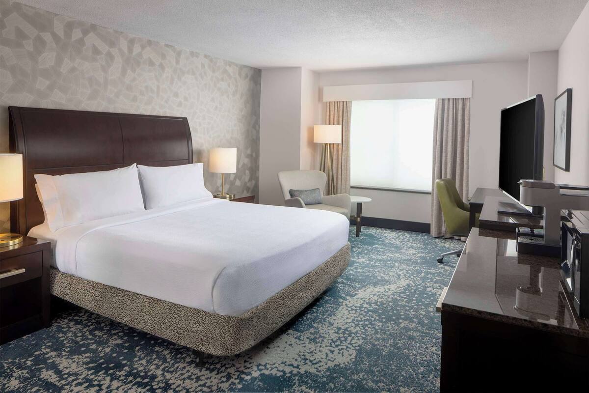 A hotel room with a large bed, a desk, a chair, a TV, and a window with curtains.