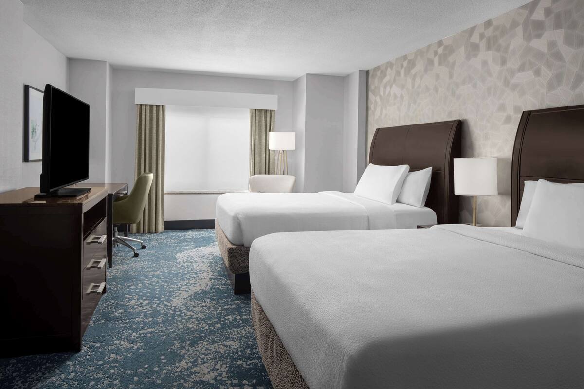 A hotel room with two beds, a desk with a television, a chair, and a window with curtains.